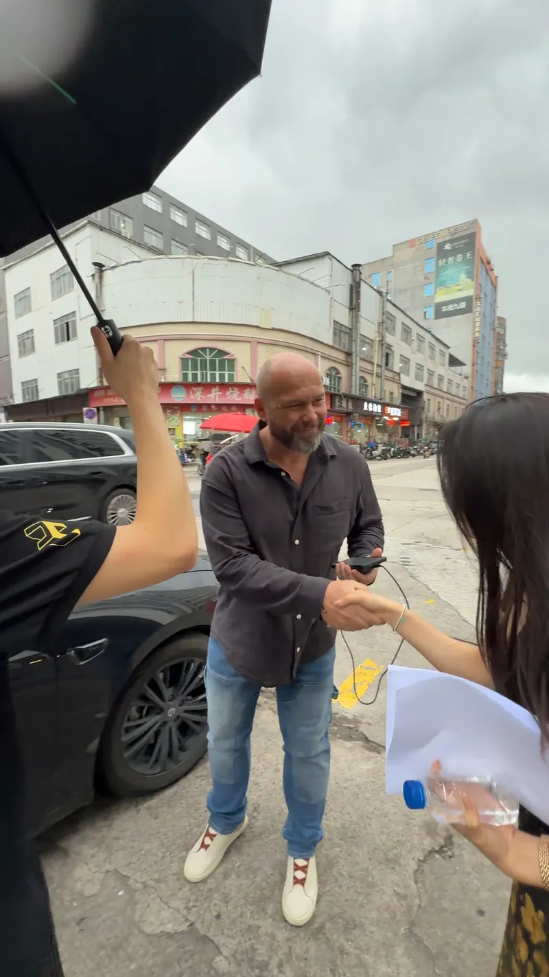 Two adults shake hands beside a parked car while another person holds an umbrella.