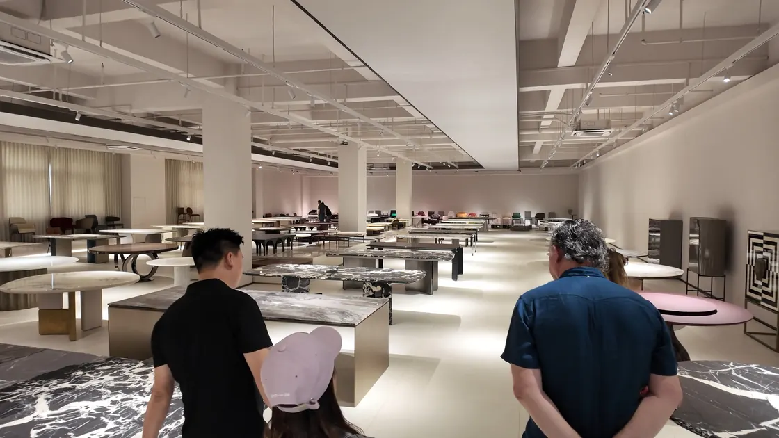 People walk into a bright showroom containing rows of round and rectangular tables.