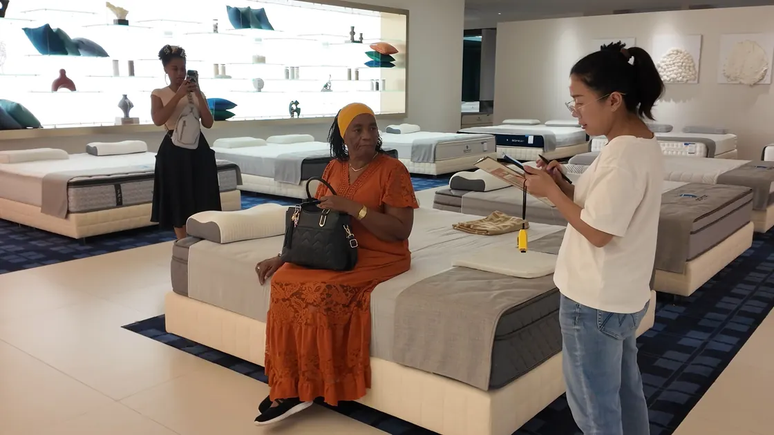 A person sits at the edge of a displayed mattress while another holds a clipboard and phone nearby.