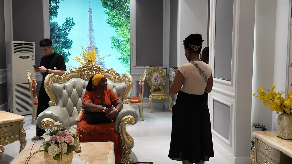A person sits in an ornate high-backed upholstered chair while another stands nearby.